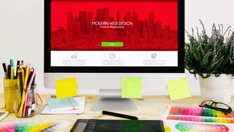 Desk with a computer showing a red website design, sticky notes and colour swatches